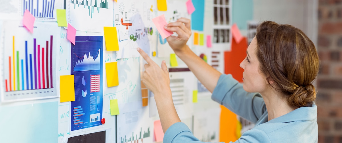 Businesswoman putting sticky notes on whiteboard a woman standing and observing a wall of charts, graphs and sticky notes